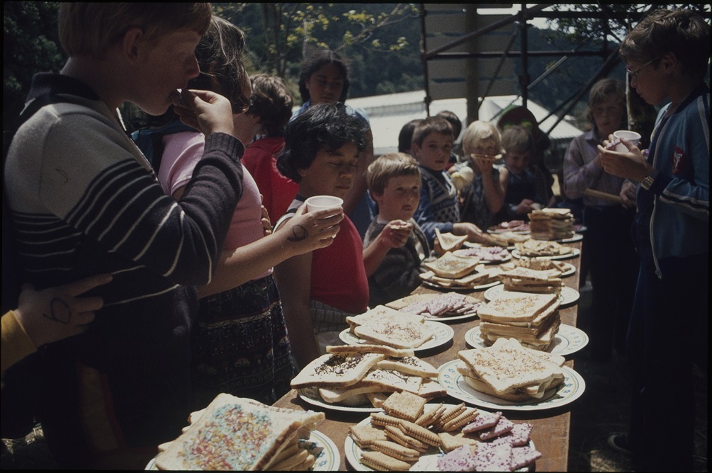 A group of children standing around a long wooden table covered in plates of fairy bread and biscuits.