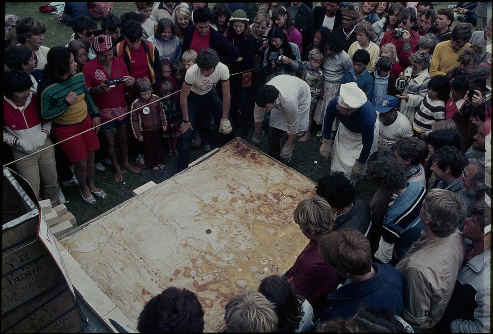 A crowd of people watch as several bakers handle a huge metre long bacon and egg pie.