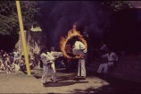 Three men in white karate uniform with black belts holding a burning hoop while a third man jumps through it.