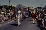 Men and women in fantastical metalic and shiny costumes walk in a street parade as families watch from the sides.