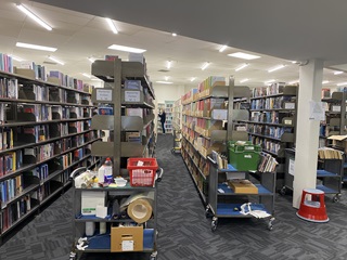 Rows of shelving all filled with books and a man looking at books deep down one of the isles, in a very large white room with florescent lighting and grey blue carpet.