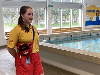 Emma Morrison standing with a brown ponytail over her right shoulder, wearing a lifeguards uniform consisting of red shorts and a yellow polo shirt with red first aid pouch slung over her shoulder, looking to her left towards a swimming pool.