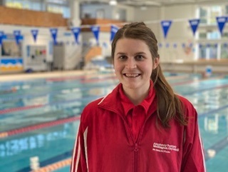 Fran Smaller smiling with a long brown ponytail wearing a red polo shirt and red jacket standing in front of a large community swimming pool with lanes.