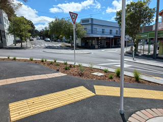 A footpath with yellow non-slip strips and a planted area next to the roadside, with a roundabout in the middle of an intersection and a two-storey building over the road with blue sky above.