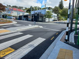 A painted pedestrian crossing with yellow non-slip strips on the footpath and a wide four-way intersection with buildings over the road and blue sky above.