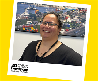 Annabelle Atkins with black hair tied up behind her head, wearing a black top and black-framed reading glasses, in front of a colourful photo of people at a city park by the ocean. Pictured in a polaroid frame with the words 20 Twenty One: Celebrating our people.