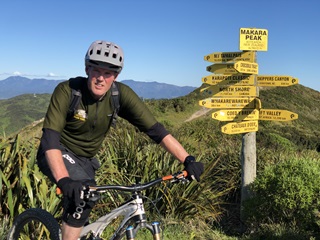 Mark Kent on his mountain bike wearing all the protective gear, perched next to a multi-directional sign post with about a dozen yellow signs pointing in different directions from the top of a hill.