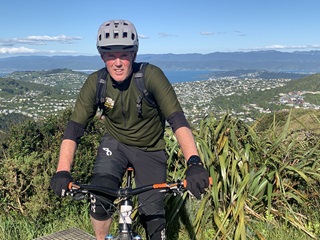Mark Kent on a mountain bike wearing all the gear a top a hill overlooking Wellington Harbour and surrounding suburbs.