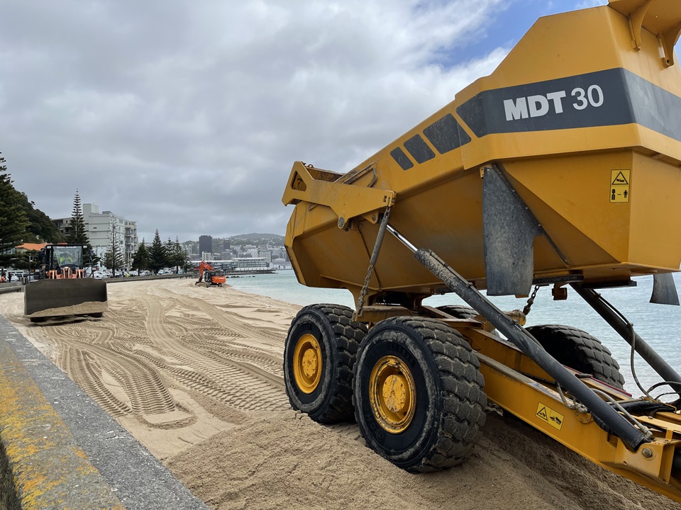 A yellow earth moving truck on Oriental Bay beach with two diggers on the sand in the background and the concrete seawall on the left.