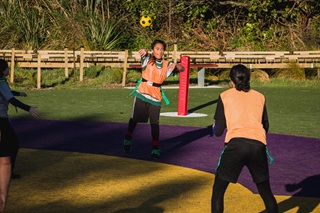 Two children wearing high-vis orange vests throwing a yellow soccer ball between them on a purple, yellow and green pitch at a sports field.