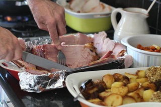 A large dish covered in foil with a ham and a person's hands slicing it up with a knife and fork, a baking dish with roast potatoes in front, and various other dishes and jugs on the table.