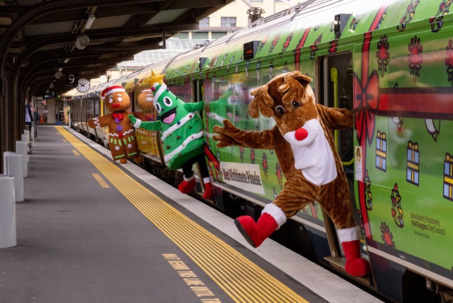 A giant fluffy reindeer, Christmas tree and gingerbread person all leaning out of the carriage doors of a train decorated with fake wrapping paper and bows.