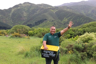 Frank Williams wearing his dark green ranger work t-shirt, holding the black #WellyWalks plastic box with yellow lid while giving a big wave and smile as he stands on a green lawn surrounded by shrubs and tall hills behind.
