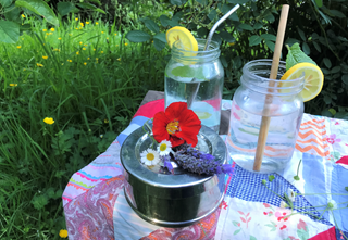 Two glass jars with slices of lemon and reusable straws, a metal food container with flowers on top, all sitting on a table covered with quilted fabric in a grassy garden.