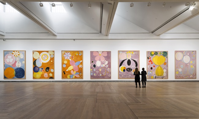 Seven very large paintings in pink, orange, yellow and blue tones, lining the wall of a gallery with the silhouettes of two onlookers admiring the art standing on a vast wooden floor.