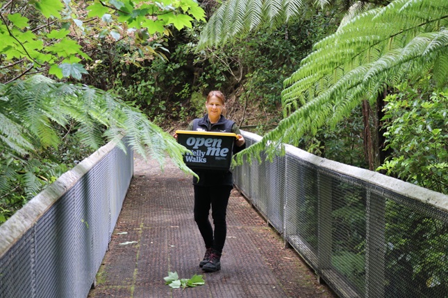 A female ranger with blond ponytail holding a large black and yellow plastic container, while standing on a narrow bridge in the middle of native bush.