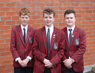 Three teenage boys wearing their red and black striped Scott's College Blazers and ties, with arms folded in front, smiling in a tidy line, standing in front of a brick wall.