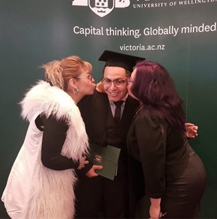 A happy young man wearing glasses and university graduation cap and gown, being kissed on either cheek by a woman with blond hair and white fluffy coat and a dark haired woman in black clothing, in front of a large dark green Vic Uni sign.