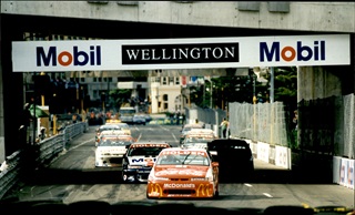A group of cars race through Wellington city under a concrete bridge which has signs that read 