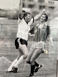 Wendi Henderson, pictured in black and white, on the soccer field wearing the gear, with her arms in the air in front of her defeated-looking opponent, after scoring her first ever goal for New Zealand  in Brisbane 1989.