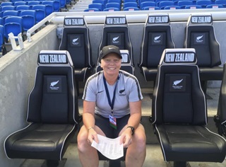 Wendi Henderson wearing a black cap, blue shorts and t-shirt and lanyard, sitting in the black New Zealand-branded seats in a stadium with blue seats behind.