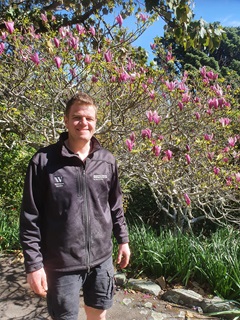 Cory smiling and standing in front of a blossoming magnolia tree in the Botanic Garden ki Paekākā.