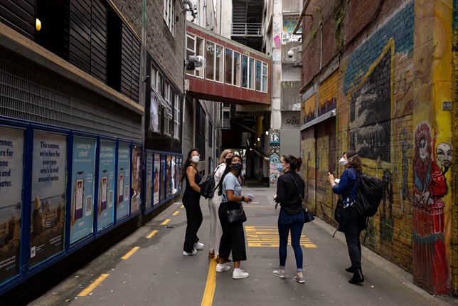 Five young woman, dressed casually and wearing masks, walking down an alleyway with posters on the left, a bright mural on the right, and a covered brown overbridge ahead.