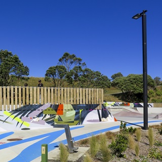A mural of bright abstract colours painted on a concrete skate park, with blue sky above and trees surrounding.