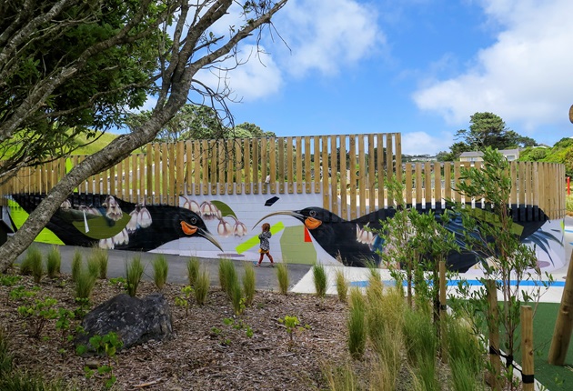 A bright mural of two huia birds looking toward one another, painted on the wall of a skateboarding park, with plantings in the foreground and blue sky above.