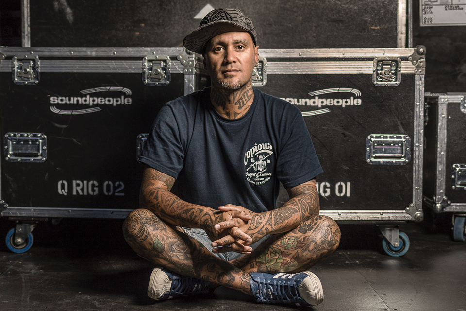 The musician Tiki Taane with tattoos on his legs and arms, wearing a dark t-shirt, shorts and a cap, sitting on the floor cross-legged in front of two large black music equipment cases.