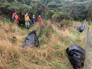 Four volunteers in brightly-coloured raincoats stand in a field of long tangled dry grass and three large black bags which are filled with weeds.