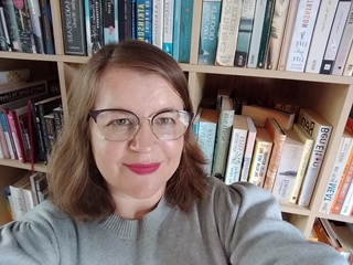 A blond woman with a friendly smile, glasses and lipstick, looking up in front of a book shelf.