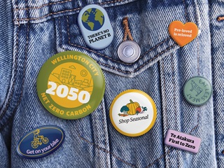The pocket of a blue denim jacket covered in seven colourful badges with various climate-themed messages, including 'eat less meat', 'shop seasonal', and 'there's no planet b'.