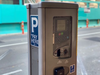 A close-up of a silver metal parking meter, with a road and bright green wall behind.