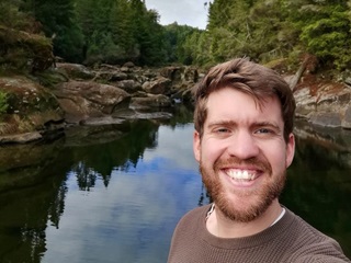 A young man with dark blond hair smiling in front of a lake with rocky surroundings and bush.
