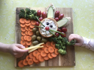 Small hands grab at items on a wooden chopping board covered in chopped up vegetable pieces and a dip, decorated to resemble a bunny rabbit. 