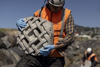 A worker in a hardhat and high-vis vest carrying a concrete tile with a woven pattern on it over rocks on a waterfront setting.