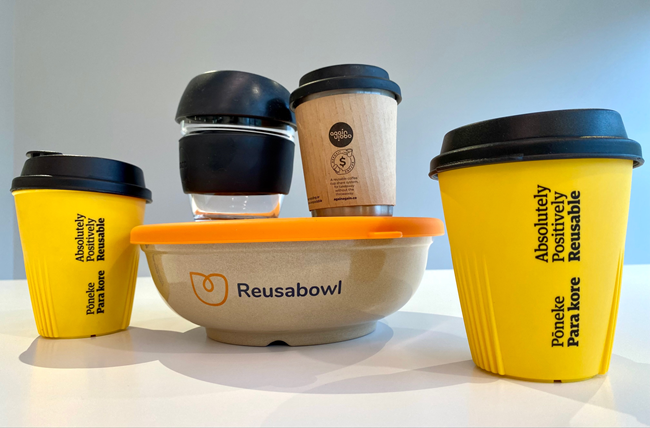 A brown reusable takeaway bowl with orange lid, with two keep cups sitting on top of it and yellow reusable coffee cups on either side.