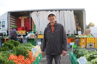 Fruit and vege stand at a market with a man standing infront of it.