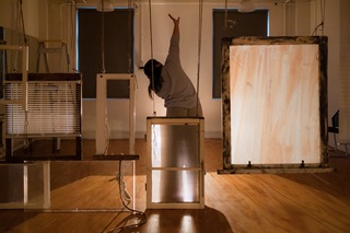 A woman mid-dance with her arm up, standing behind a set of window frames.