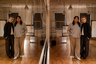 Two women standing together on the right hand side, with their reflections showing in a mirror on the left.