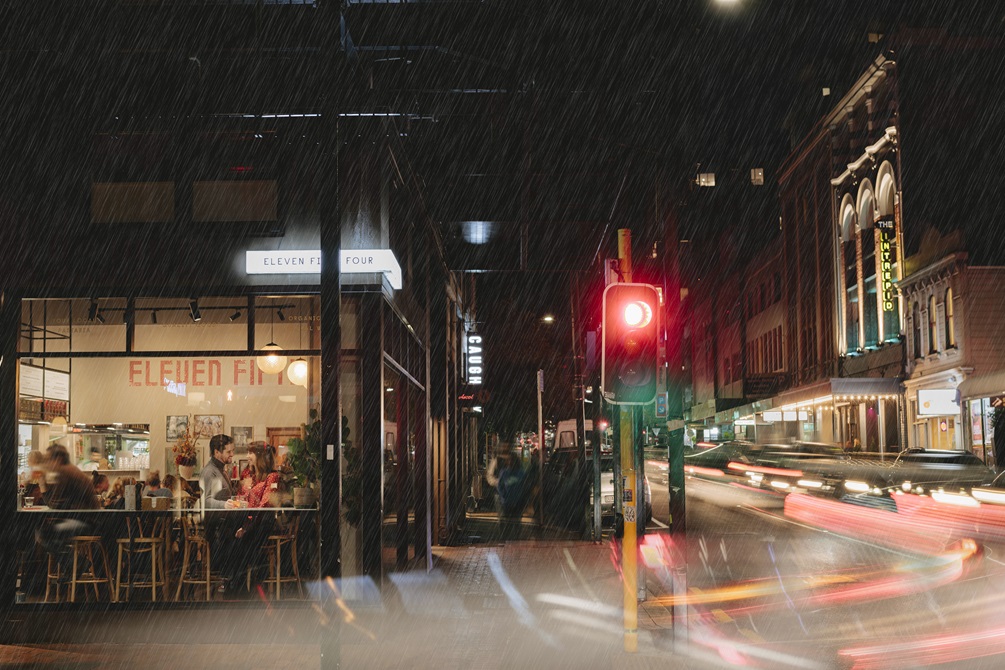 A raining evening on a city street. There is a red traffic light and vehicle headlights blur past as we look into a large window of a busy restaurant.
