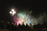 A rainbow of fireworks go off in the sky as a crowd of people look up to the dark night sky. A street light beams off to the left.