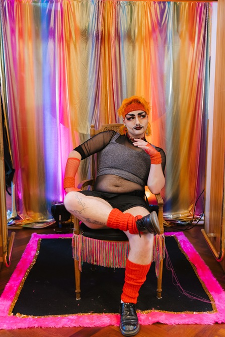A person with heavy make-up and a drawn on moustache wearing eccentric orange and black clothing, posing in a chair in front of a colourful curtain.