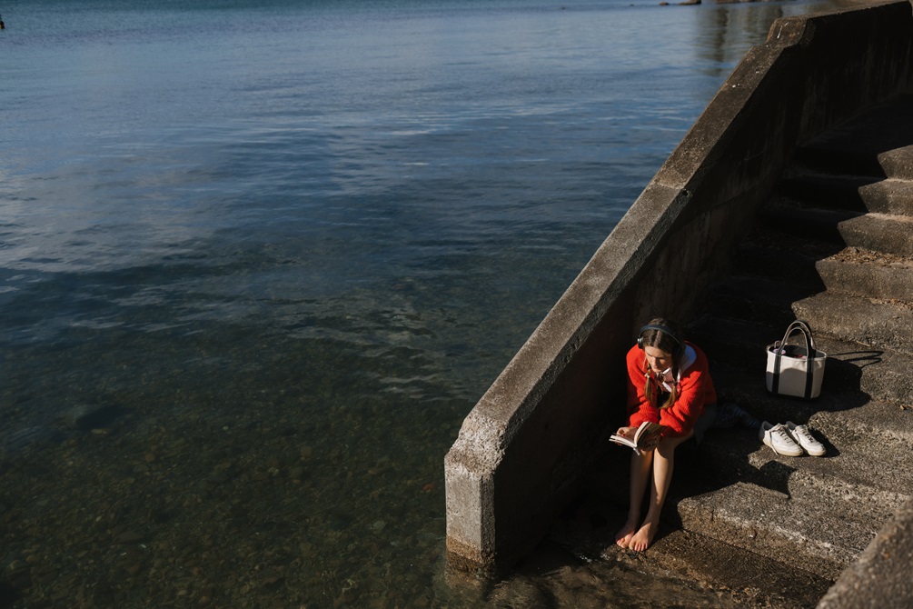 A young woman wearing a red cardigan and headphones, sitting with bare feet on concrete steps next to the ocean, as she reads a book with her belongings beside her.