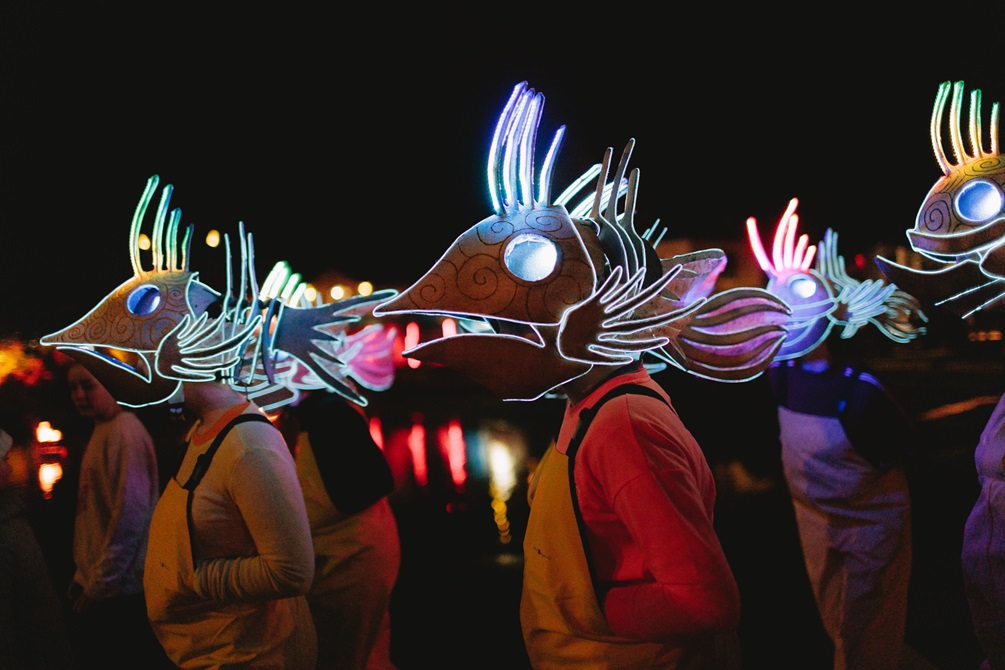 A group of people wearing giant glow-in-the-dark fish-shaped masks on their heads with a dark night sky behind them.