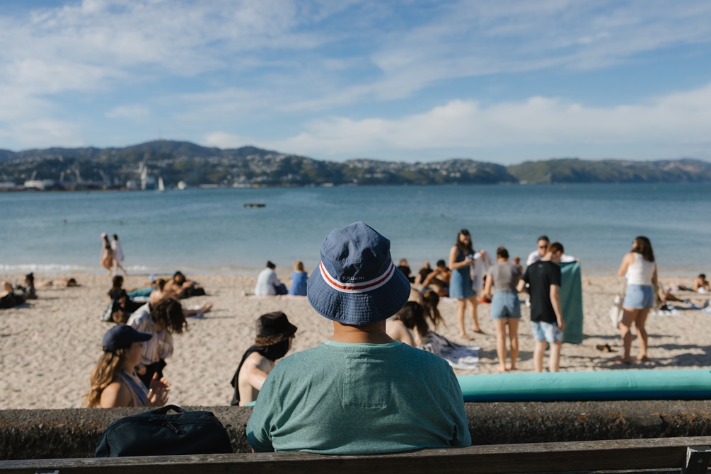 A man wearing an aqua-colour t-shirt and a blue hat sitting on a park bench, overlooking a busy beach scene, with blue skies and hills beyond.