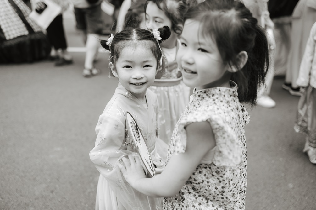 Two young girls with their hair tied in buns, wearing pretty dresses and holding each others' hands smiling. There is a crowd of people surrounding them.