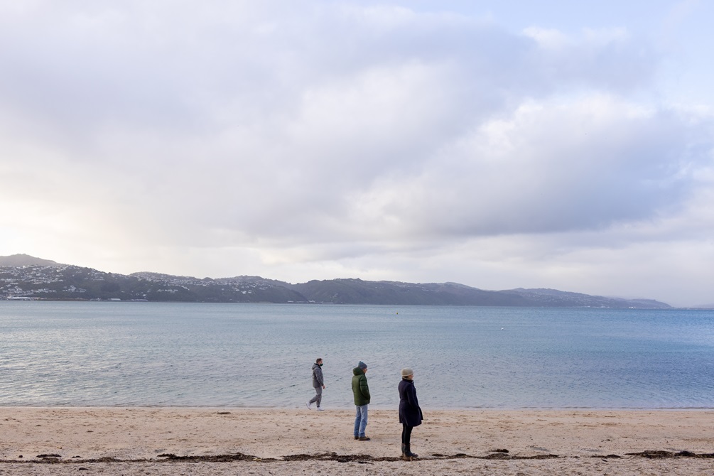 Three people standing  on the beach in a line with a few metres separating them. The water behind them is still and you can see hills in the distance.