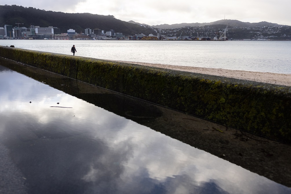 A puddle in the foreground that reflects the clouds in the sky. A beach in the background with a person walking in the distance.